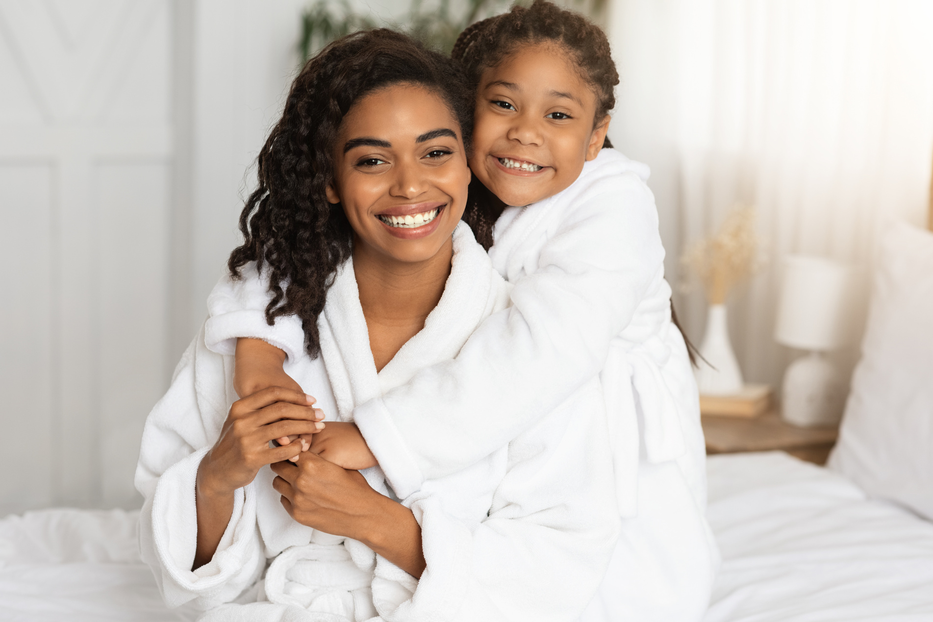 Beautiful Happy Black Mom And Daughter Posing On Bed In White Bathrobes